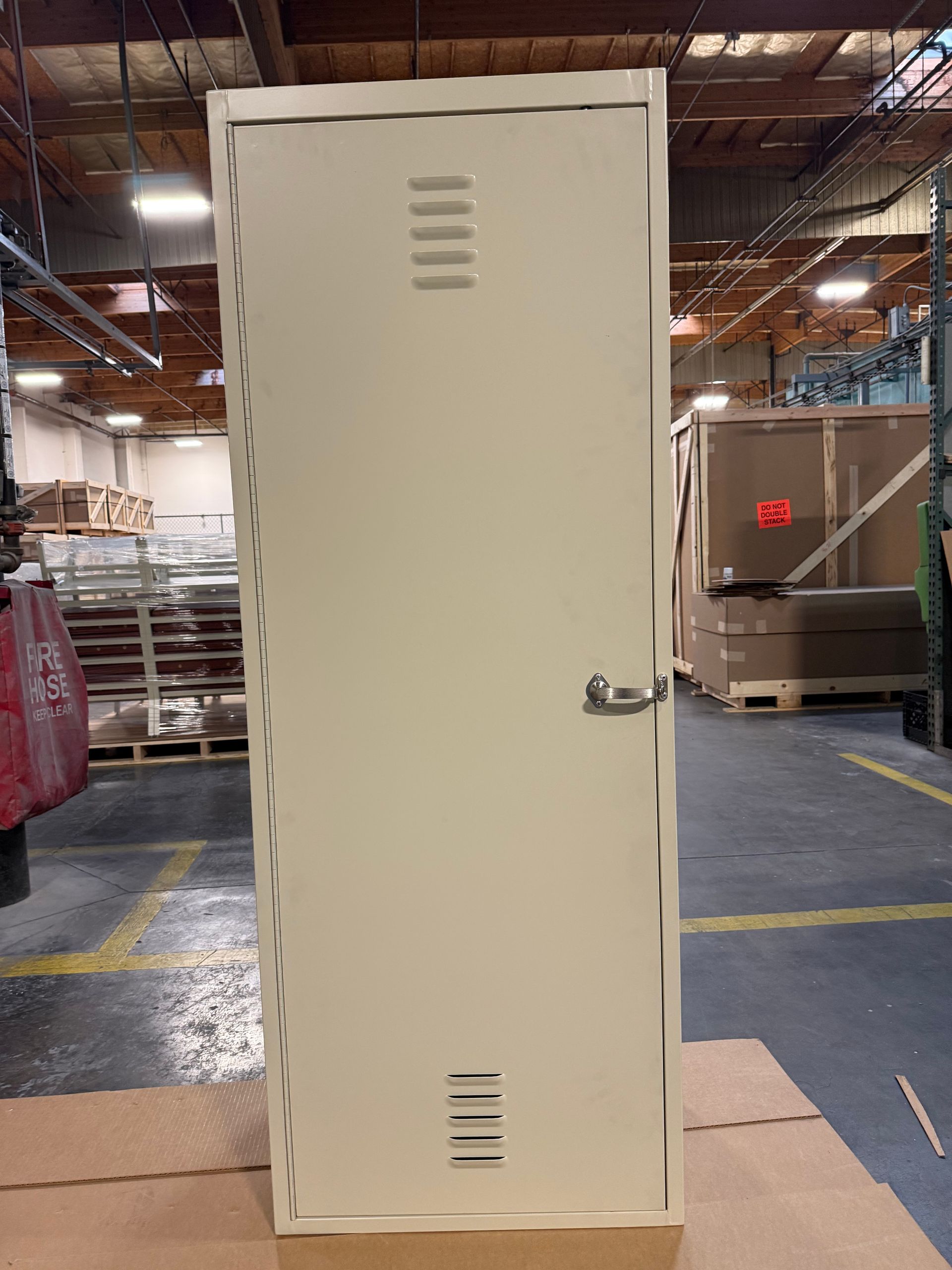 Tan metal locker in an industrial setting, door closed with ventilation slots and a handle.