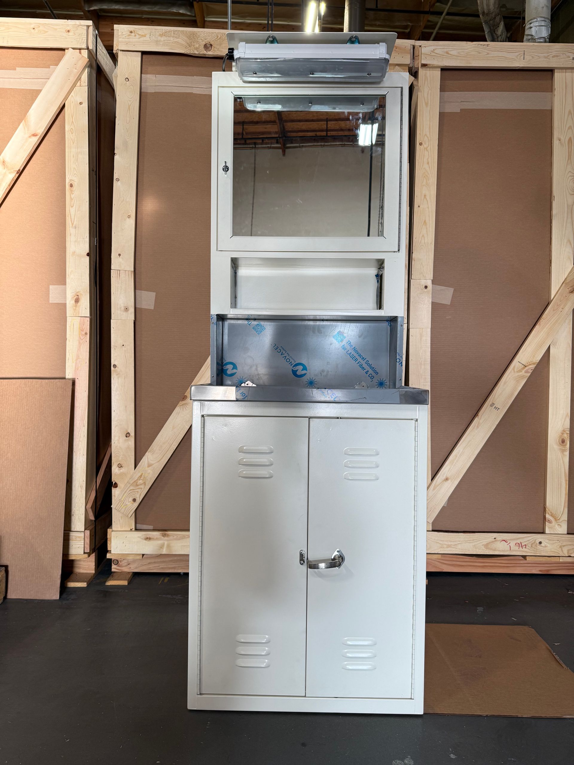 White metal medical handwashing station with mirror and cabinet, set against wooden crates.