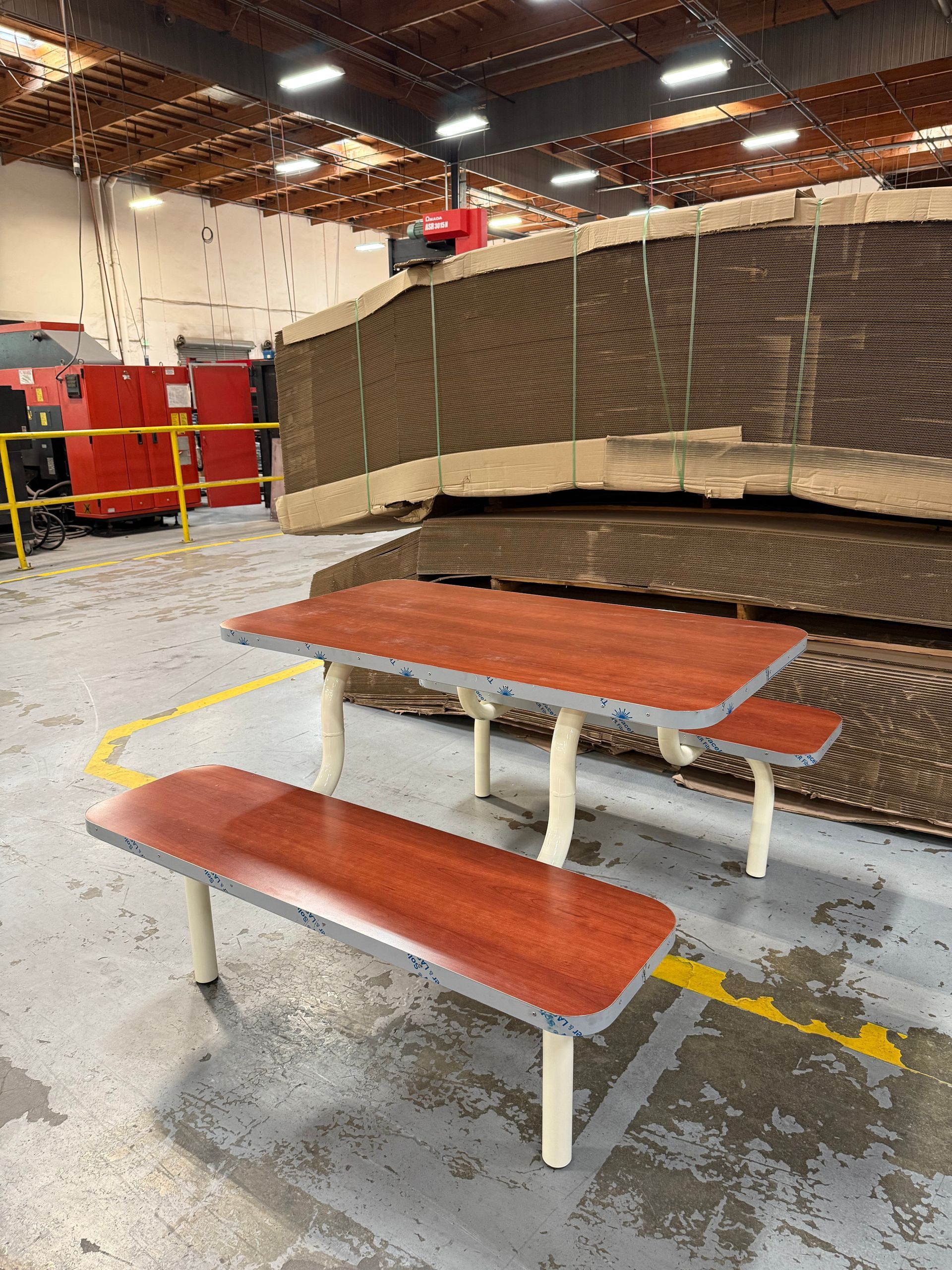 Picnic table and bench in a warehouse. Brown tabletop, white legs. Cardboard structure in the background.