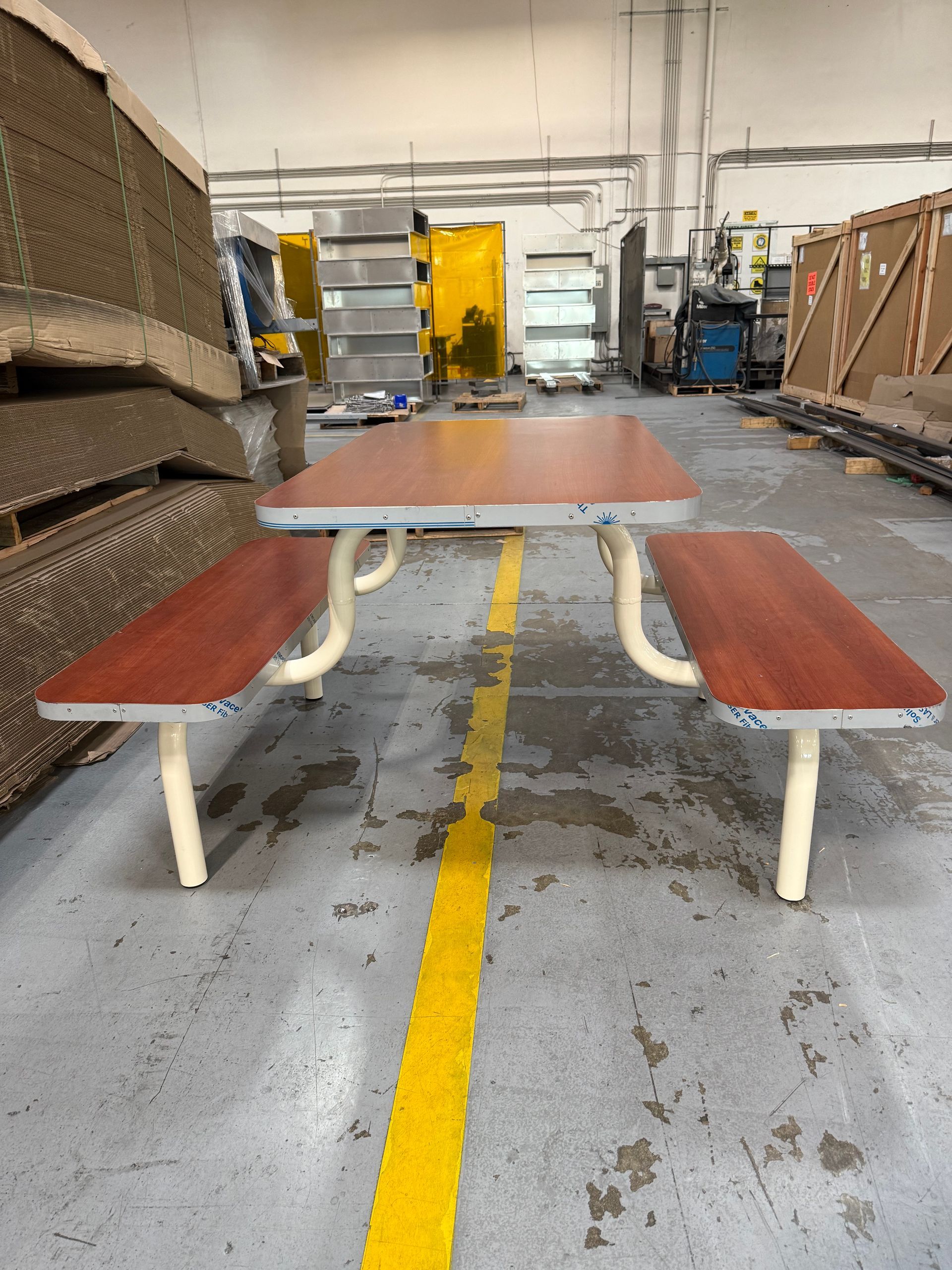 Picnic table with red-brown tabletop and benches, beige legs, on a gray concrete floor in a warehouse setting.