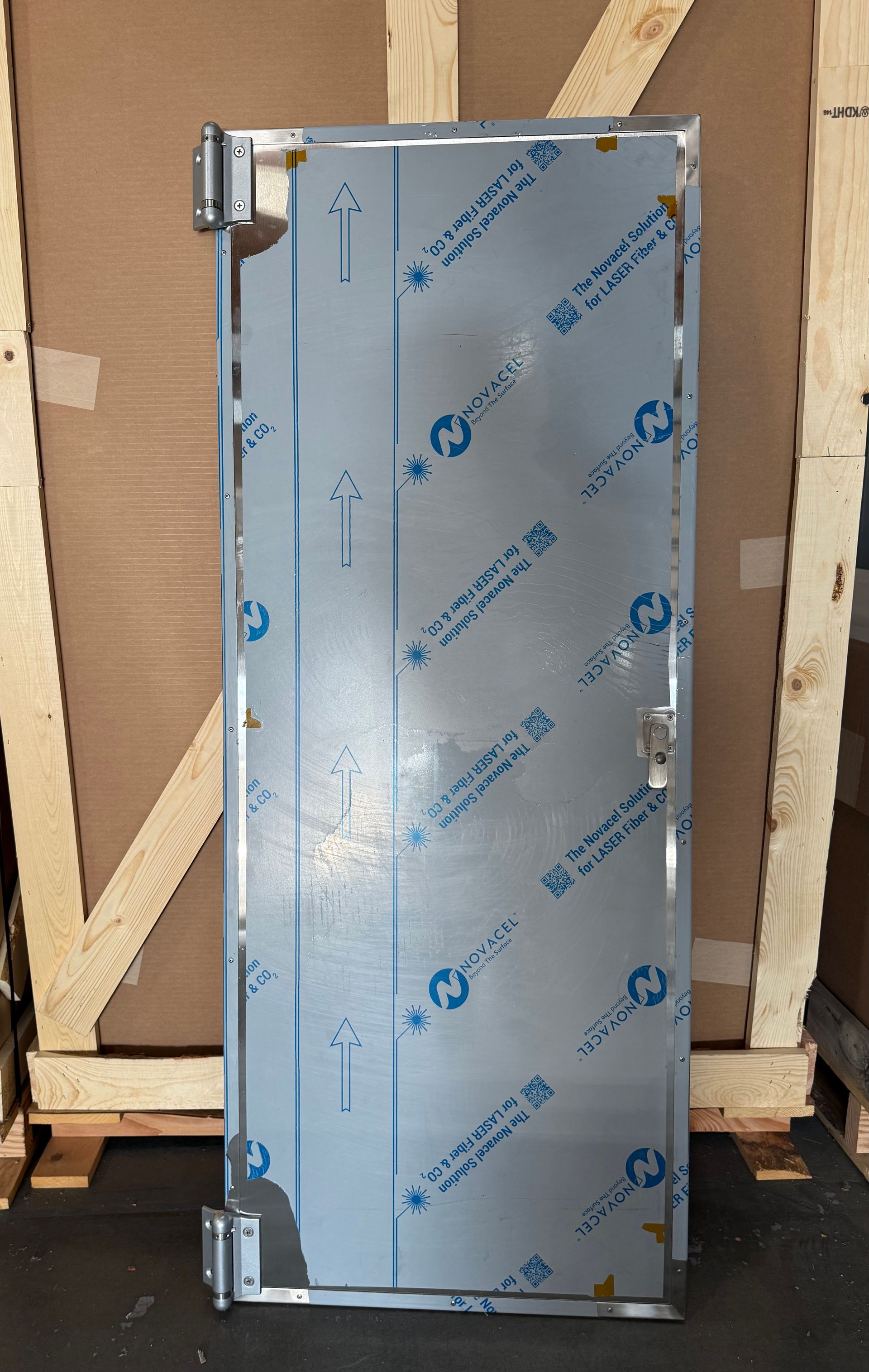 Stainless steel door with protective film, hinges, and handle, leaning against a wooden crate.