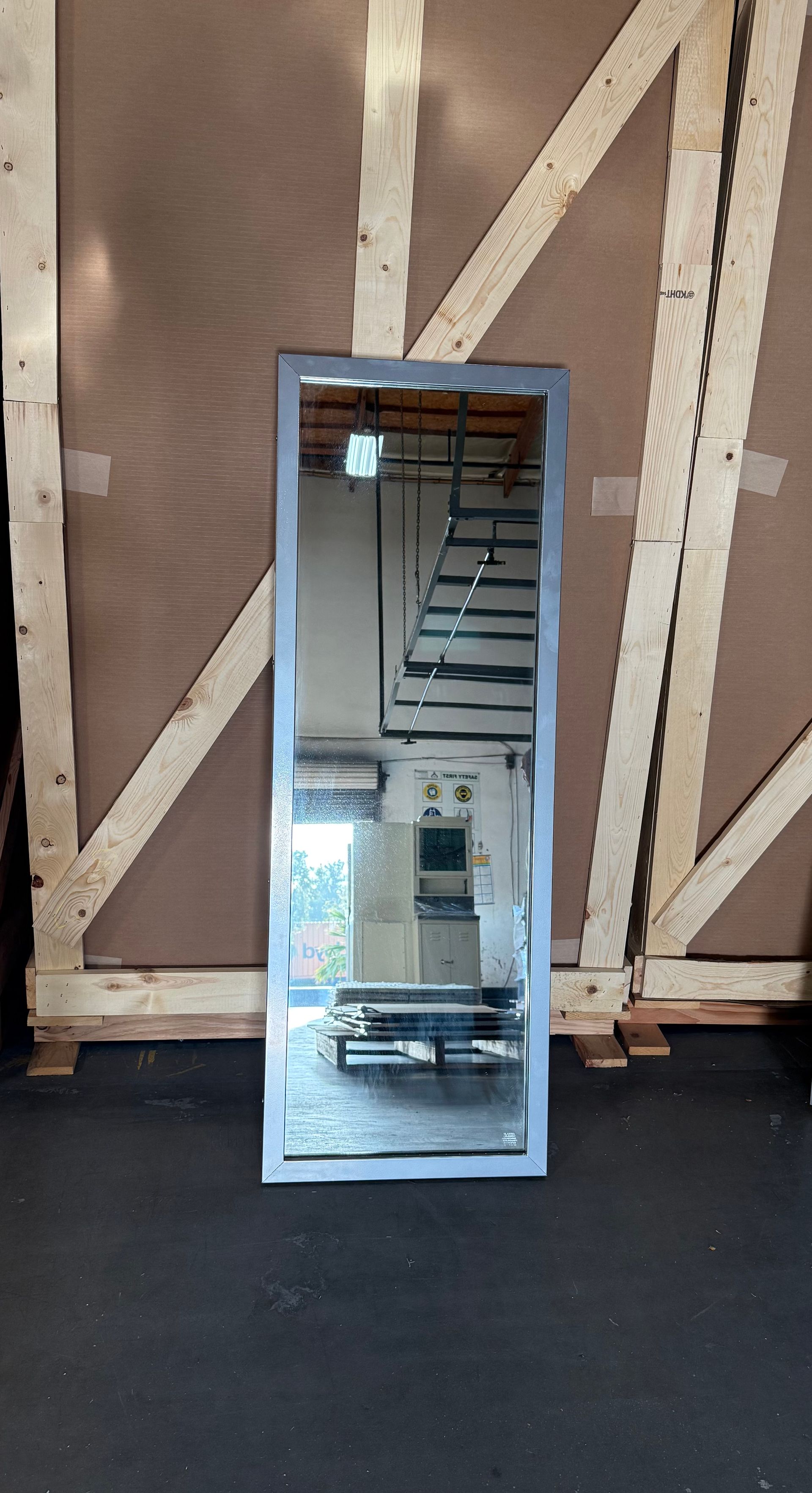 Full-length rectangular mirror with a silver frame, leaning against a wooden backdrop.