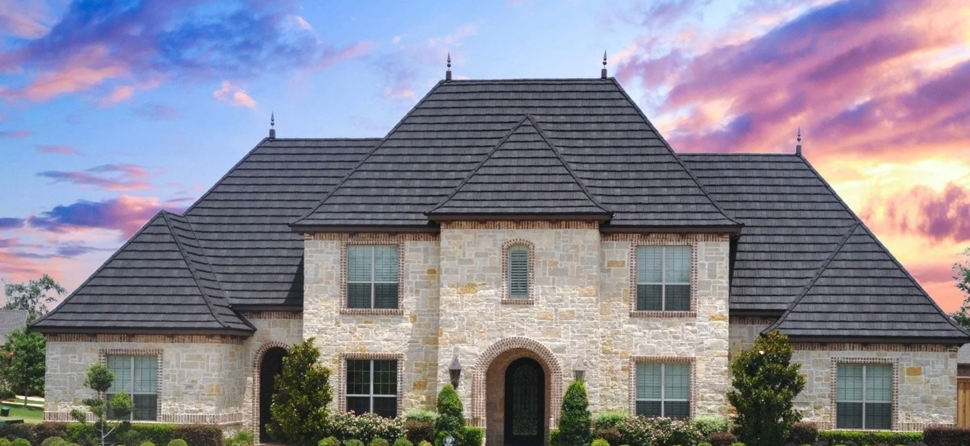 A large brick house with a black roof and a sunset in the background.