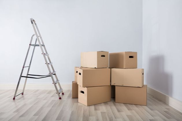 A stepladder stands beside a stack of cardboard moving boxes in a corner of an empty room with light wood flooring.