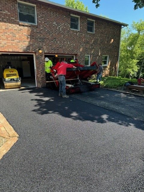 A group of people are working on a driveway in front of a brick house.