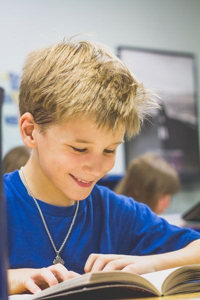 A young boy in a blue shirt is reading a book in a classroom.