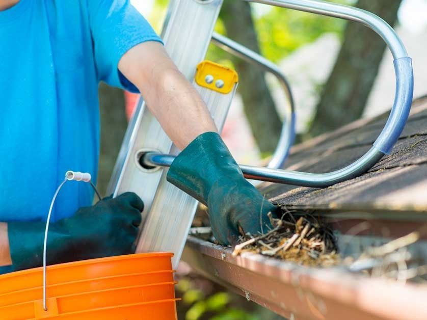 Worker in blue shirt fitting gutters with a metal ladder and orange bucket nearby