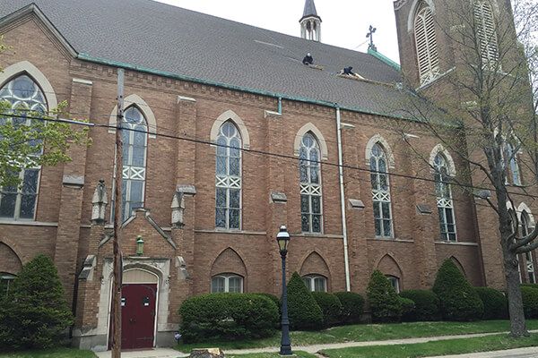 Brick church building with tall arched windows, red door, and a stone bell tower amid trees and shrubs