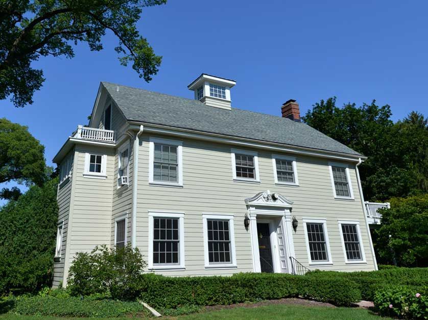White two-story house with black shutters and a small cupola, surrounded by trees and shrubs under a blue sky