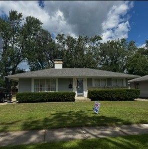 Single-story white house with green shutters, front lawn, trees, and a “For Sale” sign.