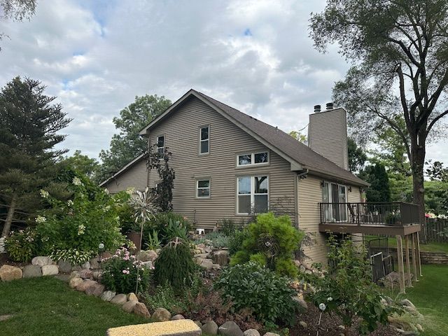 Tan house with stone chimney, deck, and lush garden under a cloudy sky