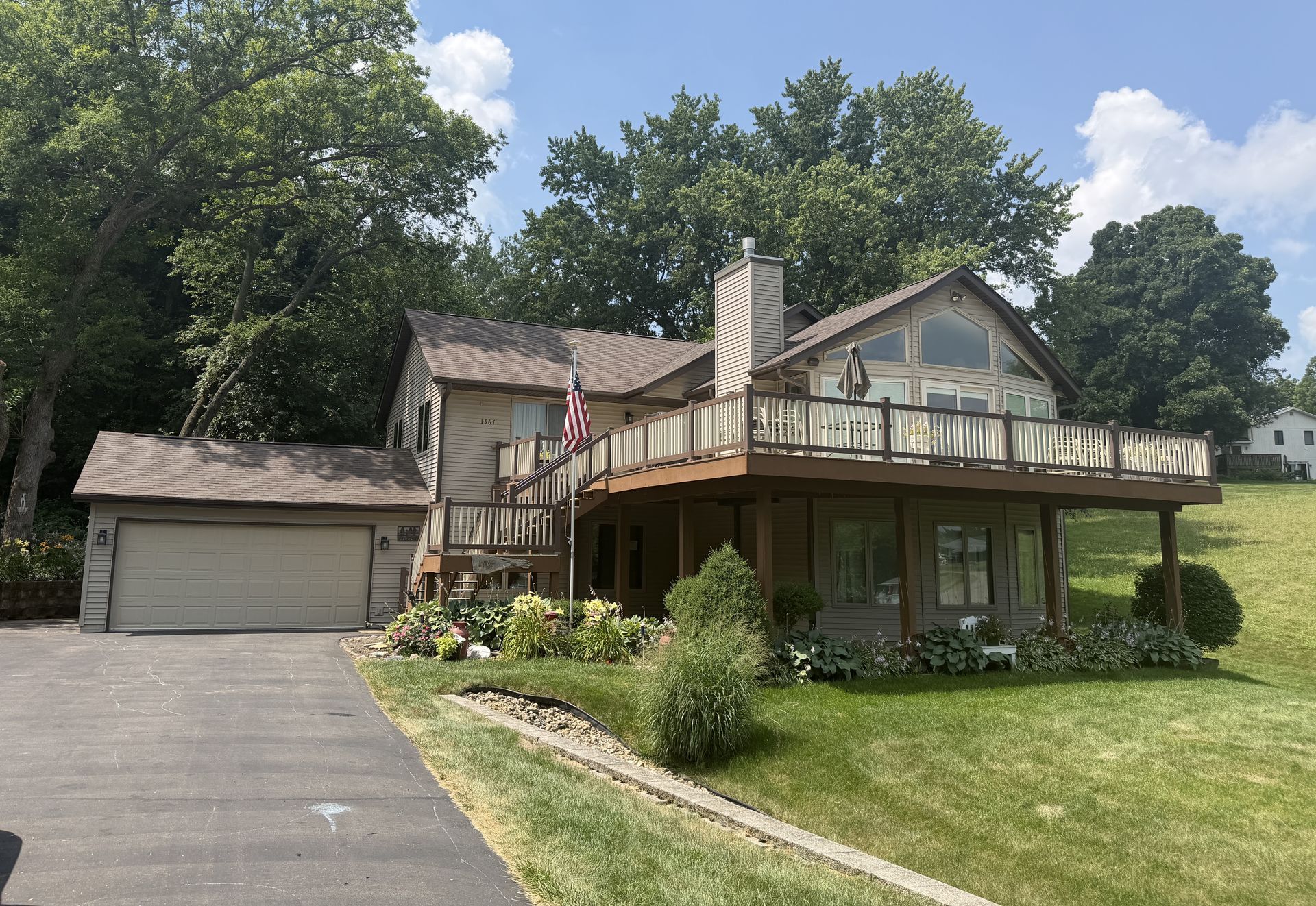 Suburban house with a large wraparound deck, attached garage, driveway, and green lawn under a blue sky