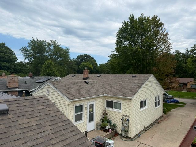 Exterior view of a beige house with a gray shingled roof and small patio, surrounded by trees.