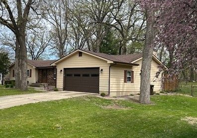 Single-story beige house with a two-car garage and a front lawn, framed by tall trees.