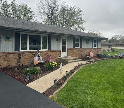 Single-story brick house with a front walkway, flower beds, and a green lawn under a cloudy sky