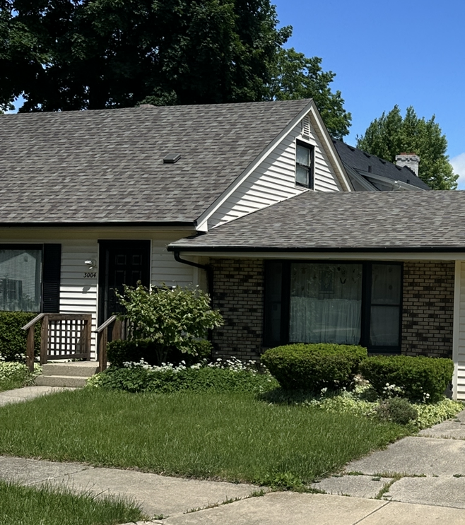 Suburban house with gray roof, front lawn, shrubs, and a small porch under a clear blue sky