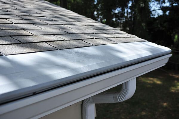 Gutter along the edge of a shingled roof on a house, with a downspout below.