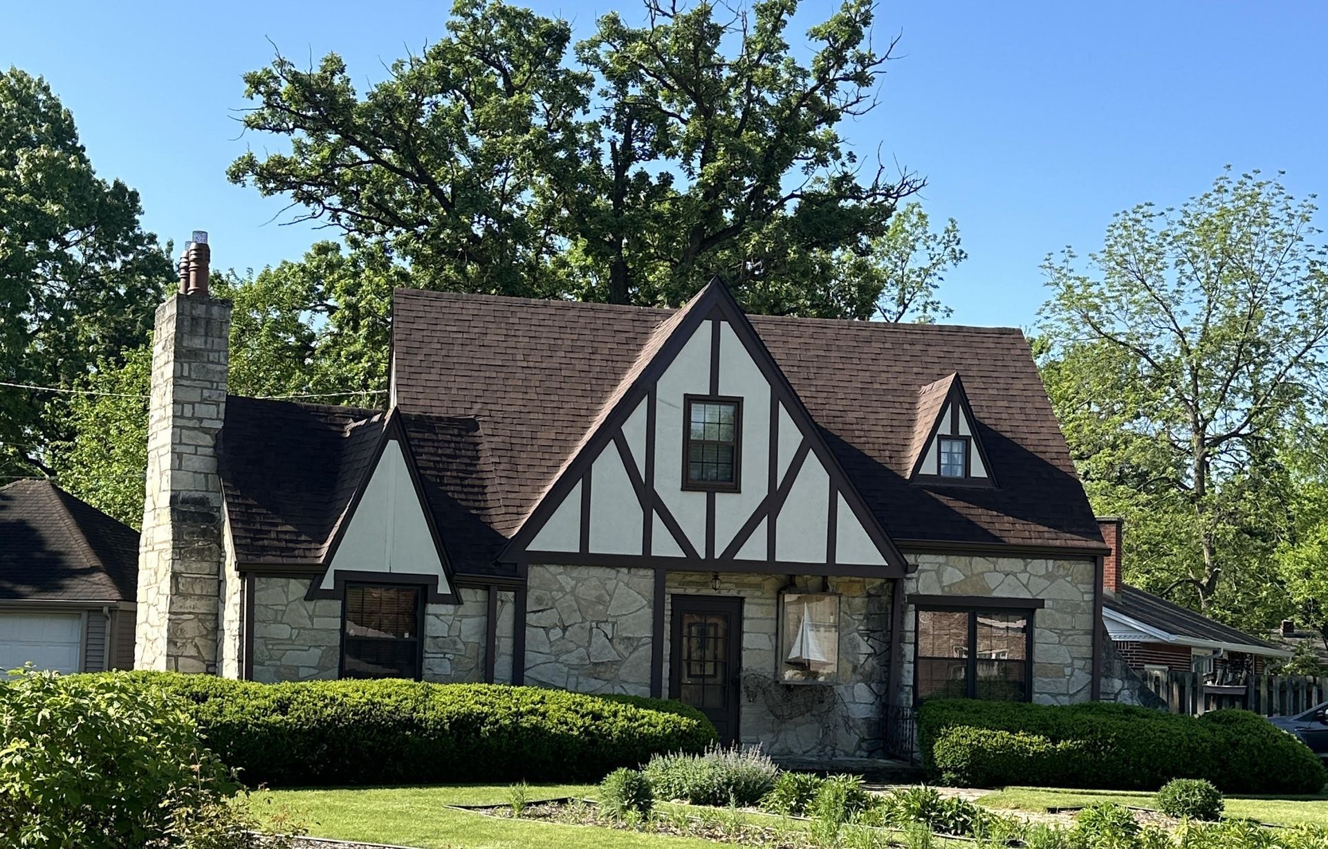 Tudor-style stone house with dark brown roof, trimmed hedges, and tall trees in a sunny yard