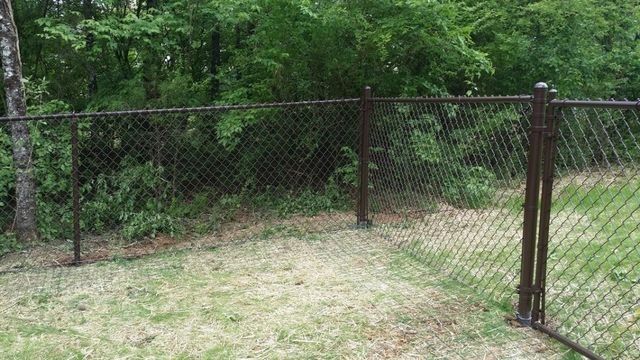 Brown chain-link fence in grassy yard with trees in the background.