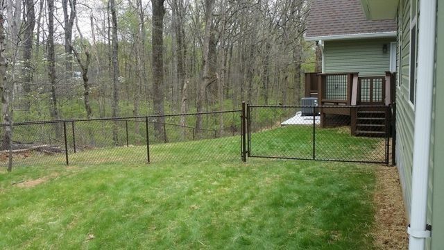 Lawn with a black fence leading to a house with a wooden deck and wooded background.