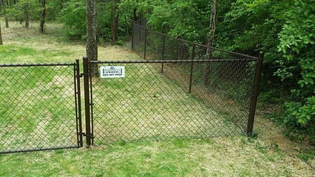 Dark chain-link fence with a gate encloses a grassy area near trees. A sign is attached to the fence.