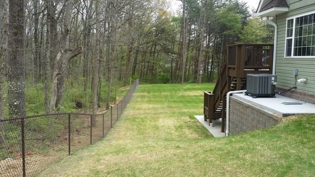 Brown chain-link fence lines a grassy yard, with a home and trees in the background.