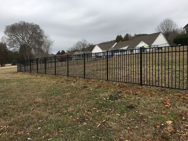 Black metal fence along a grassy slope, with houses in the background under a cloudy sky.