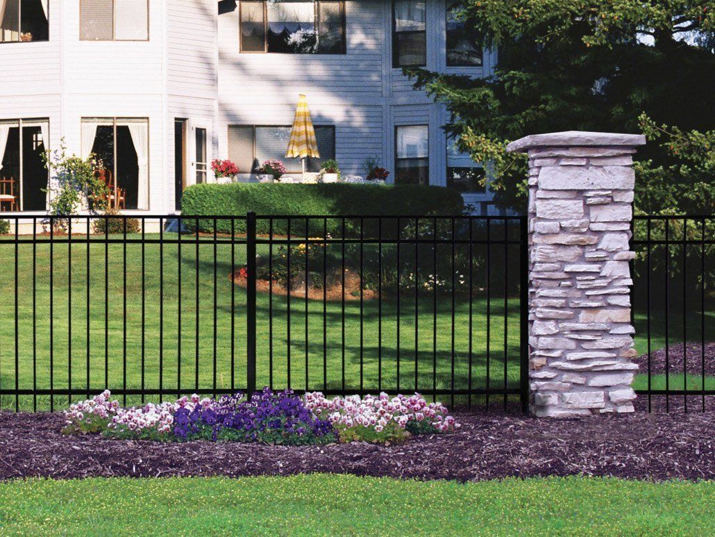 Black metal fence with stone pillar, in front of a house, purple flowers in the foreground.