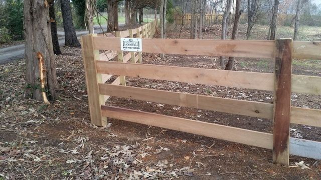 Wooden split-rail fence in a wooded area, with a sign and a dirt ground.