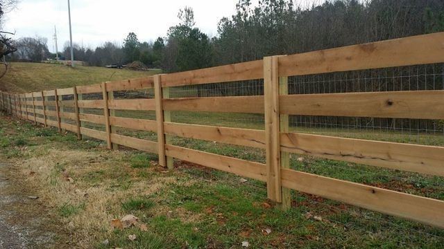 Wooden fence with three horizontal rails, metal mesh, and posts in a grassy field.