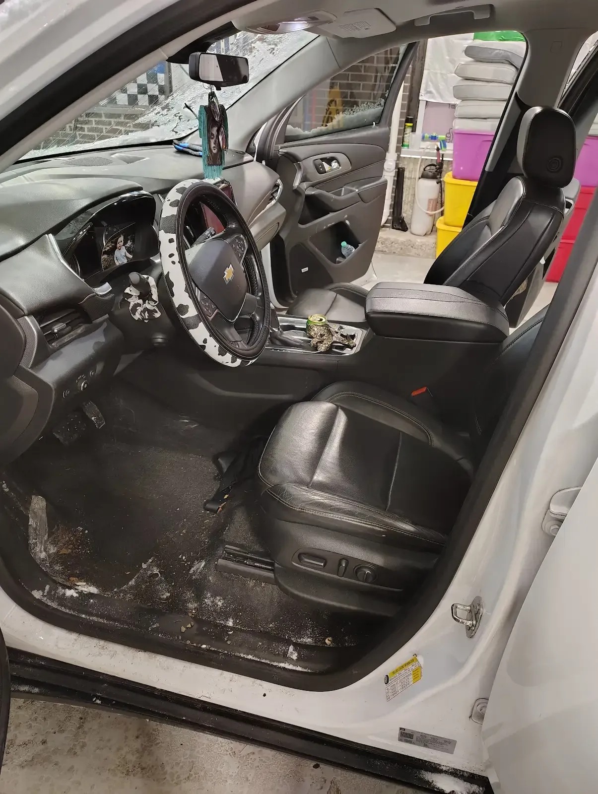 The interior of a white car featuring black leather seats, a patterned steering wheel cover, and a messy floor mat.