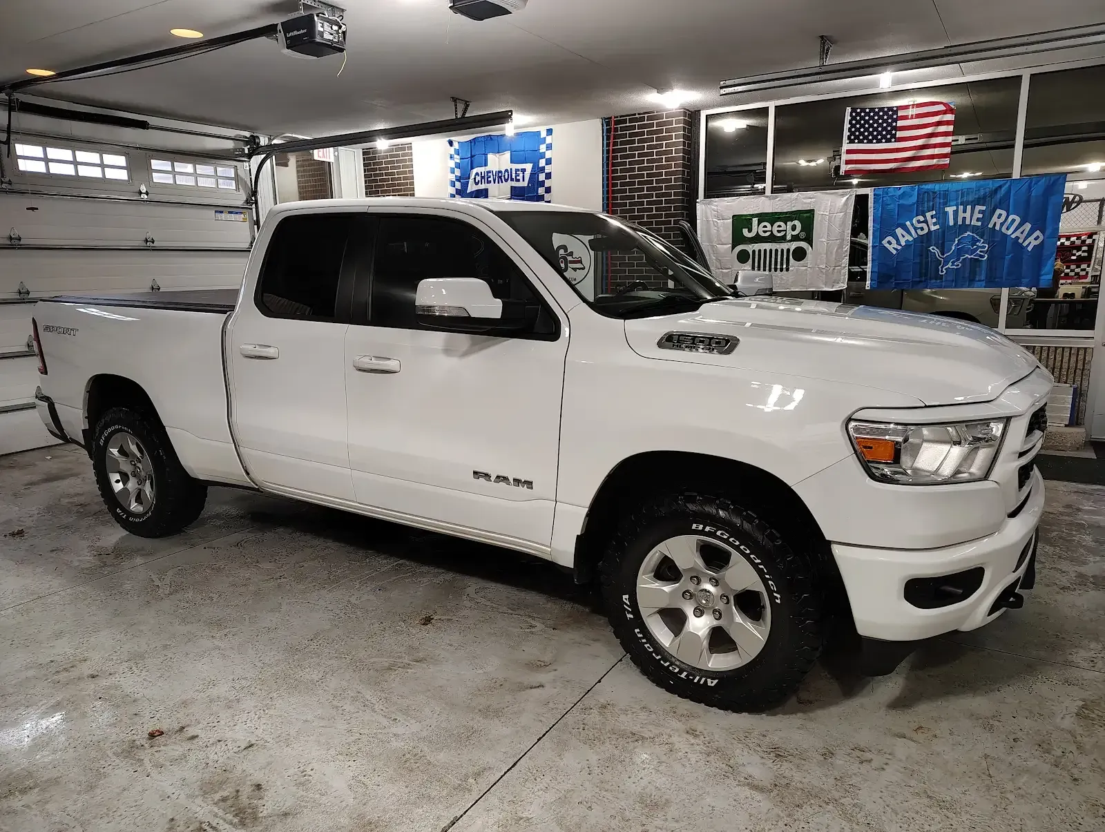 A white Ram 1500 pickup truck parked inside a well-lit garage with American and Jeep flags hanging on the back wall.