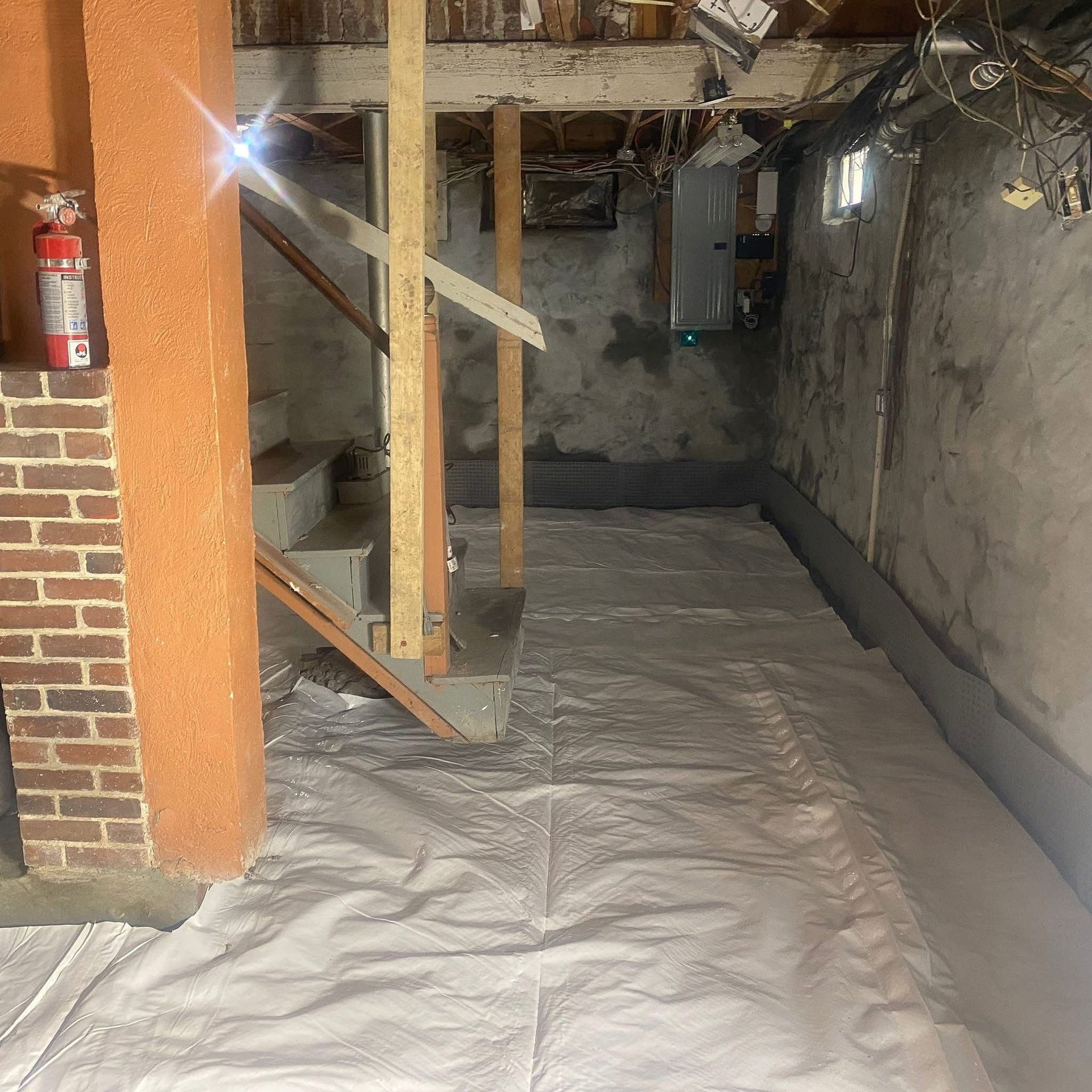 Basement with white ground cover, brick pillar, wooden stairs, and concrete walls. A fire extinguisher is visible.
