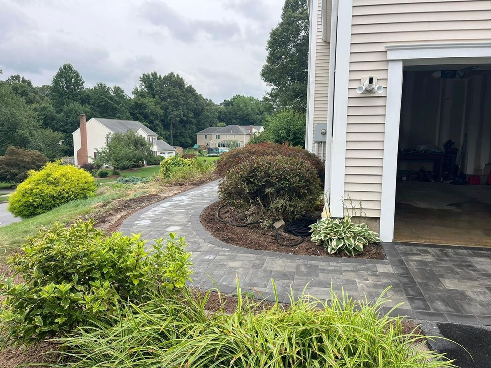 Paver walkway curving towards a garage, edged by shrubs and greenery