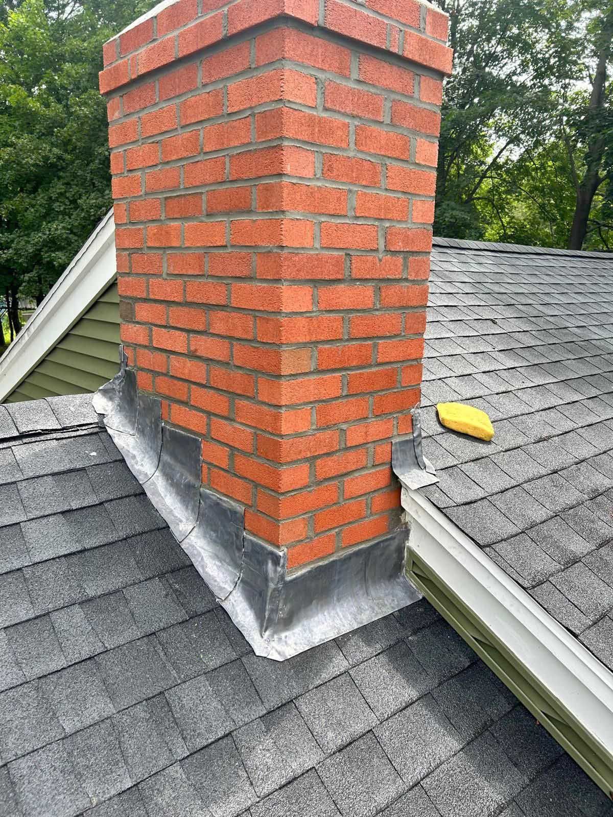 Brick chimney on a dark shingled roof with flashing, and a yellow object.