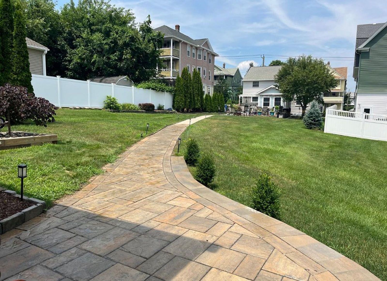 Brick pathway winds through a grassy backyard, surrounded by a white fence and houses