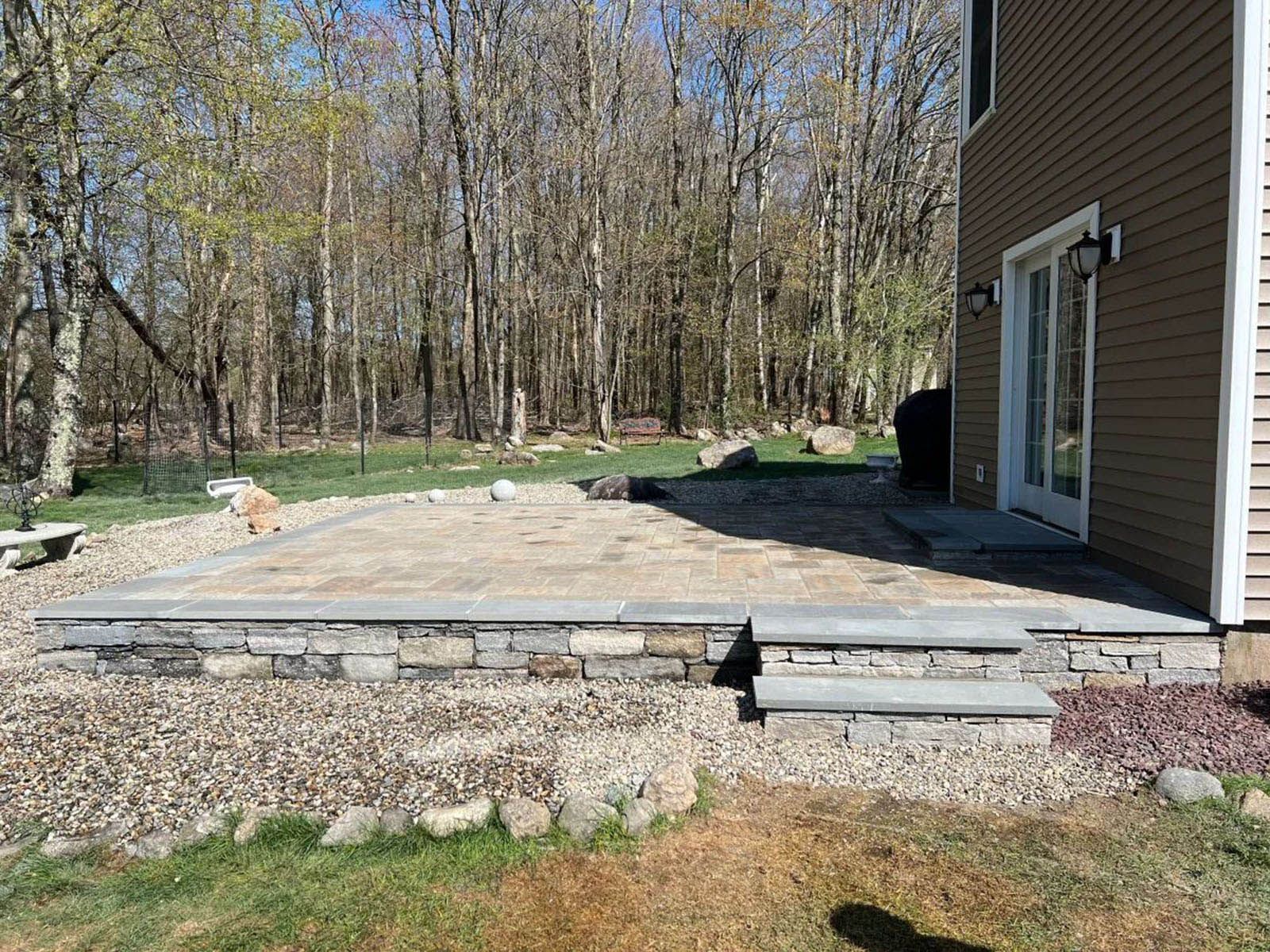 Stone patio with steps and retaining wall next to a house, gravel ground, trees in the background