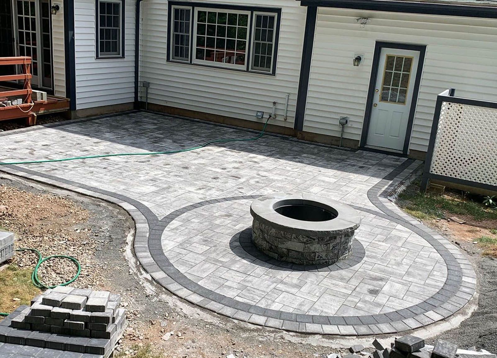 Brick patio with fire pit, bordered in gray and black pavers, adjacent to a white house