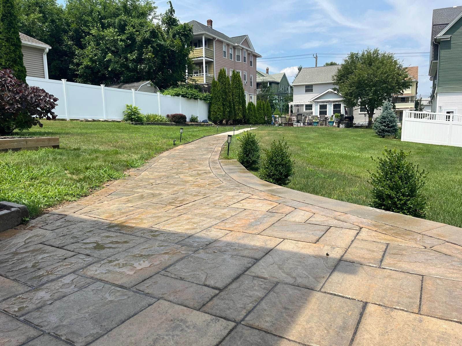 Stone patio and walkway leading through a grassy yard toward houses under a blue sky