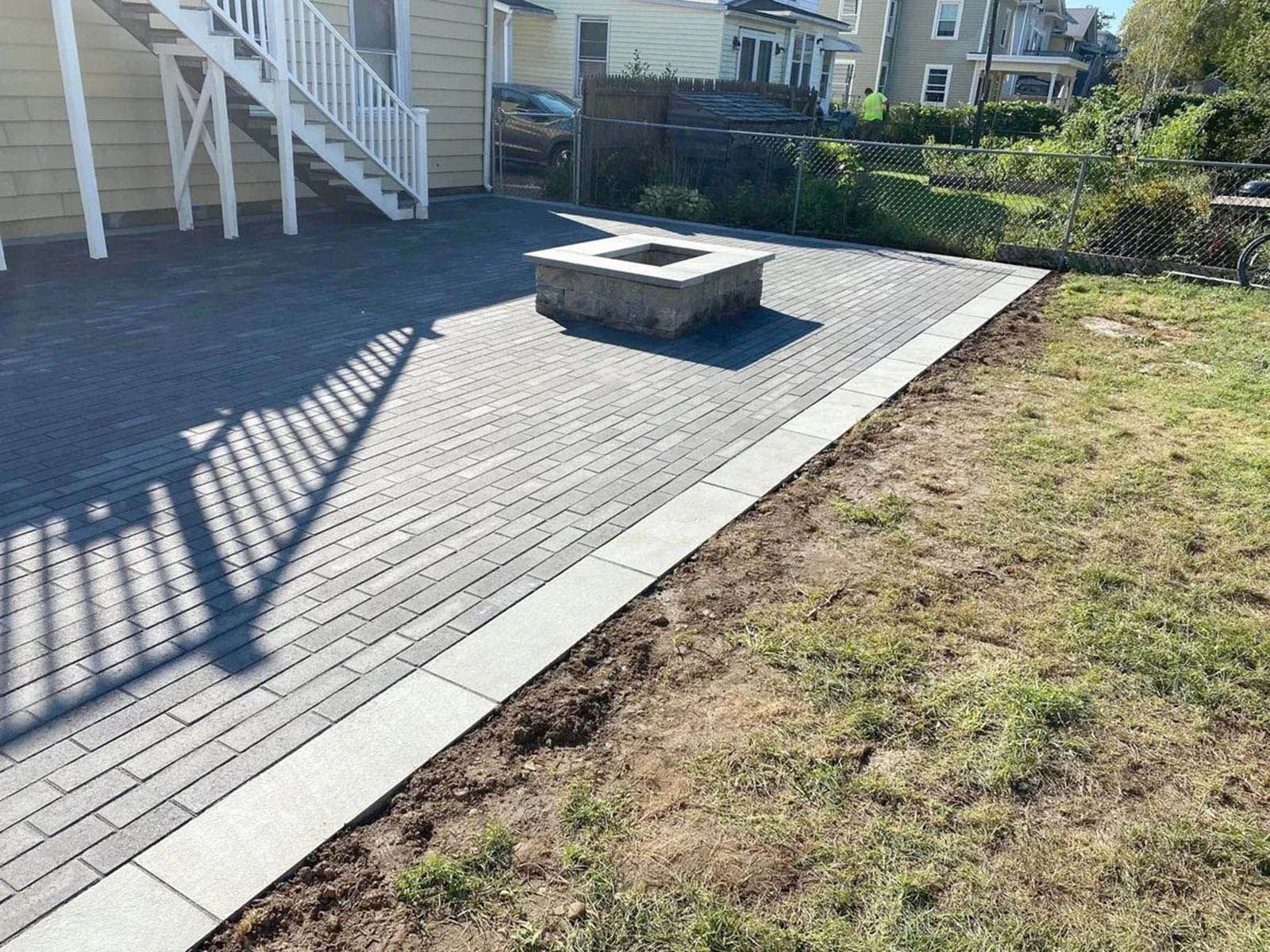 Brick patio with fire pit, bordered by concrete and grass, adjacent to a house with stairs