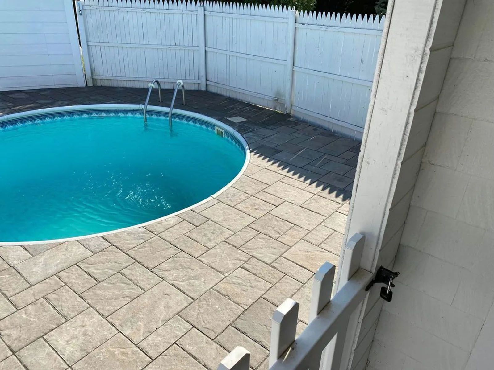 Above-ground pool surrounded by gray pavers and a white picket fence