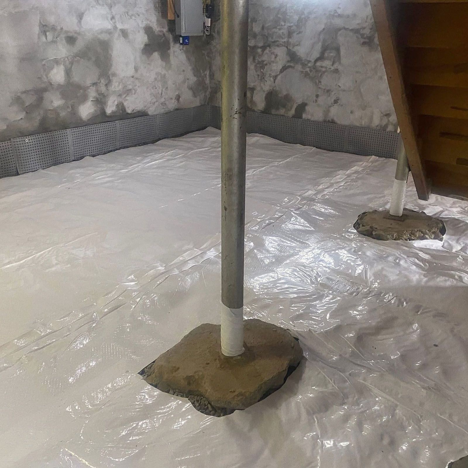 A basement interior with white plastic sheeting on the floor, concrete walls, and support poles
