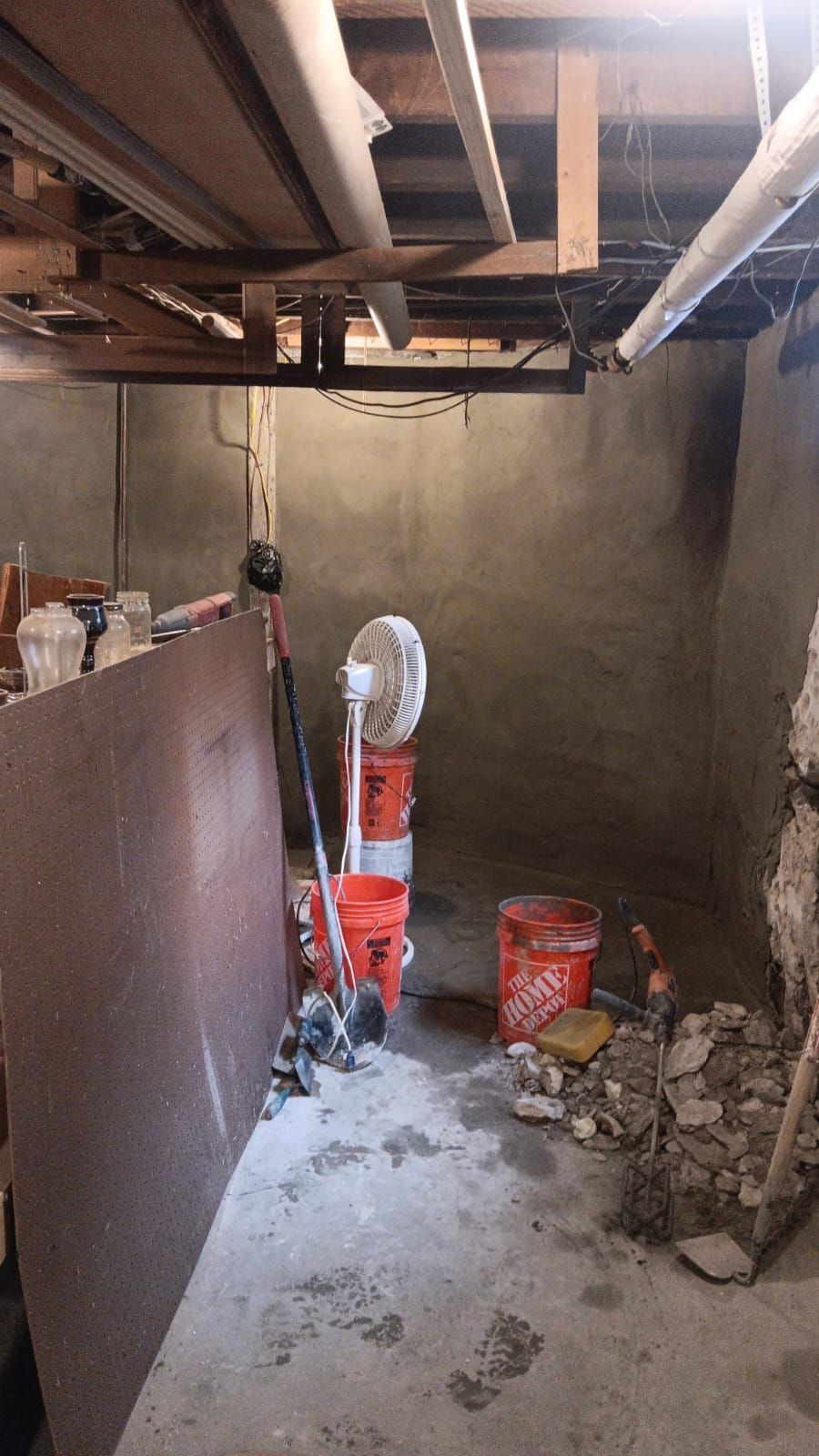 Basement renovation: concrete walls, exposed pipes, construction debris, fan on buckets, plywood leans against wall.