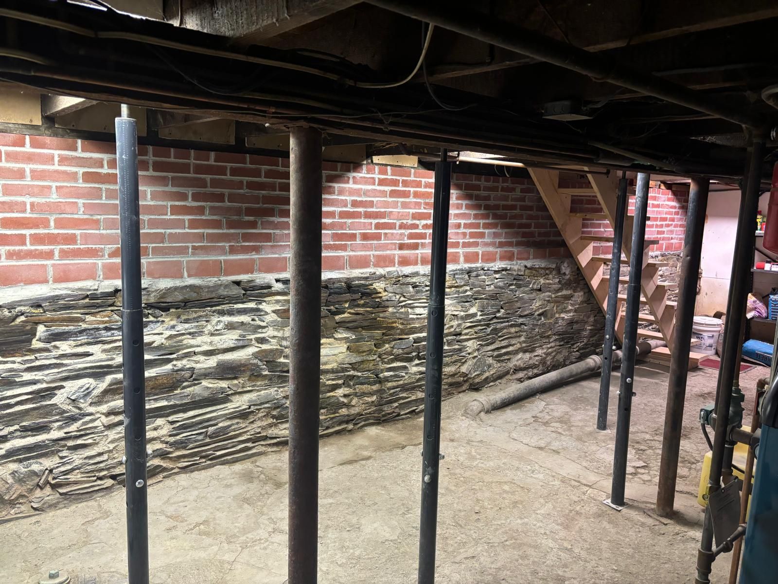 Basement interior with support columns, stone foundation, brick wall, and wooden stairs.