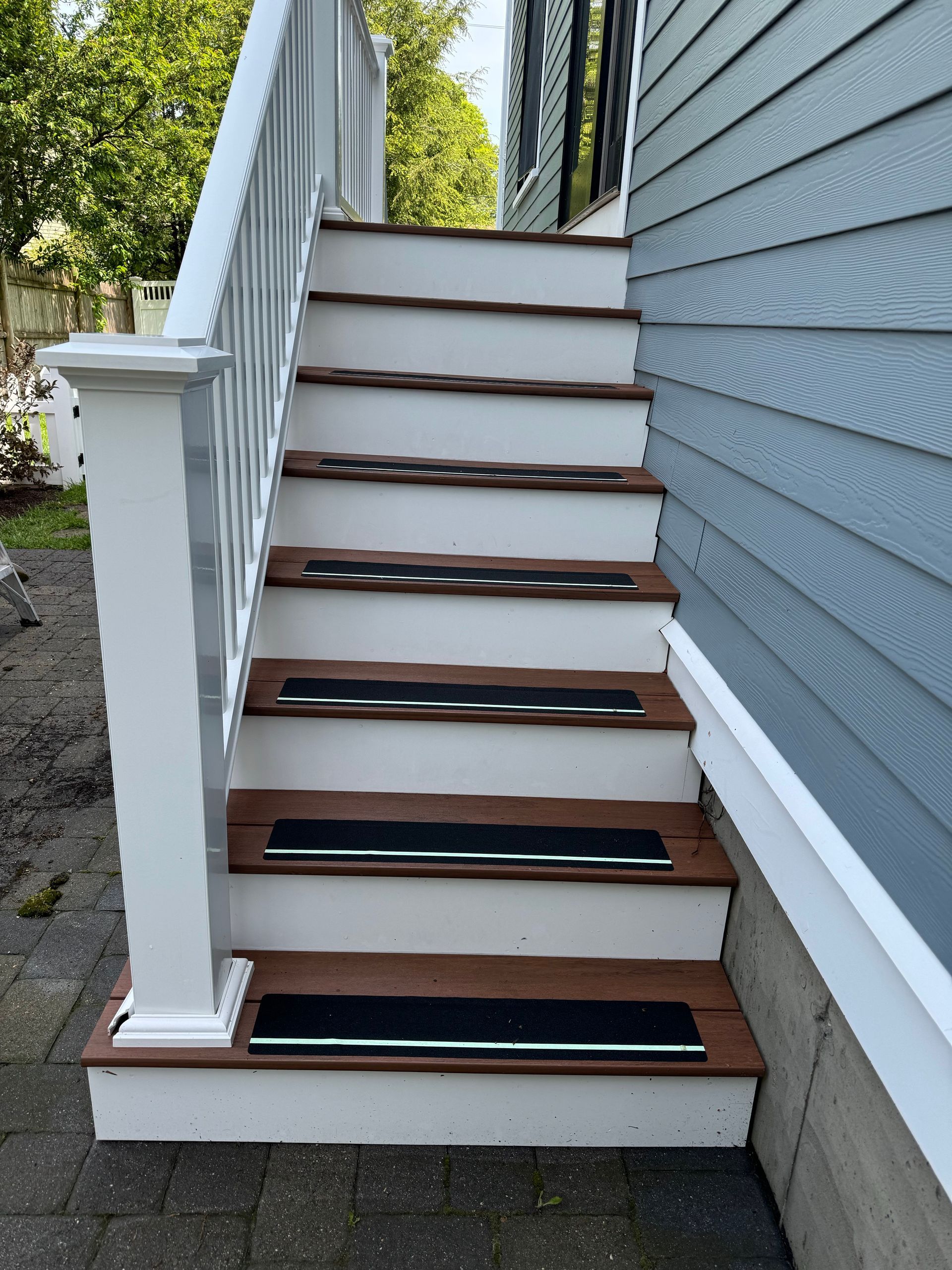Outdoor wooden staircase with white risers, brown treads, and black anti-slip strips. Blue siding and white railing.