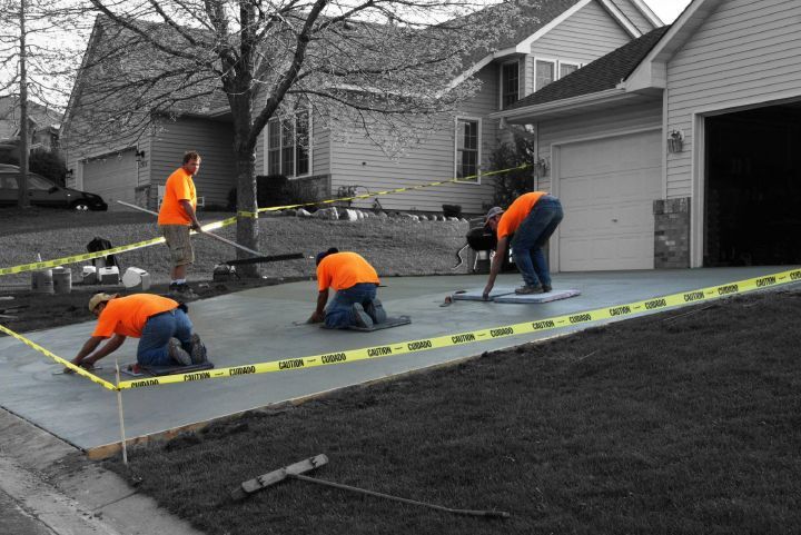 Four workers in orange shirts smooth a fresh concrete driveway outside a suburban house behind caution tape.