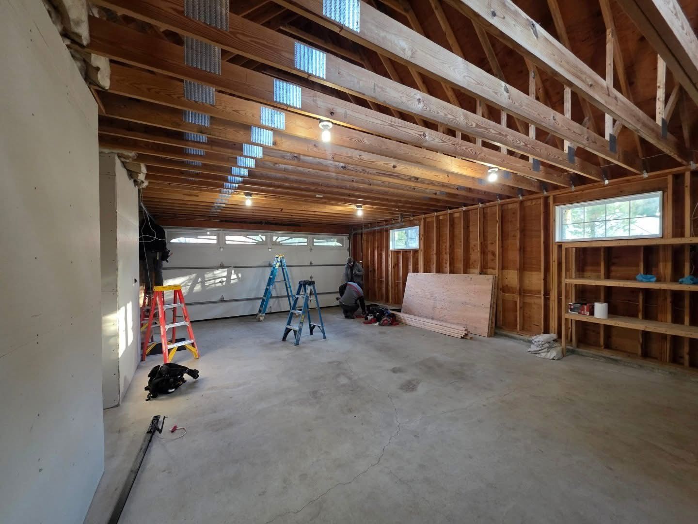 Garage interior under construction: exposed framing, concrete floor, insulation, two windows, tools and a ladder visible.