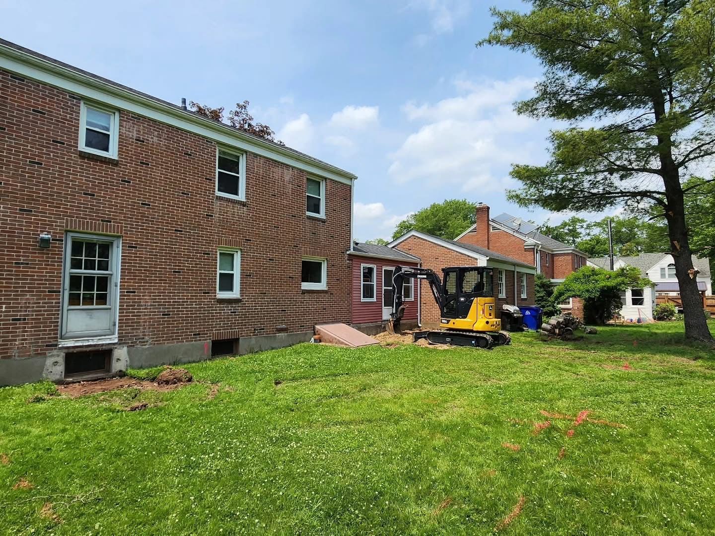 Backyard with brick house, small excavator, and other houses visible under a blue sky.