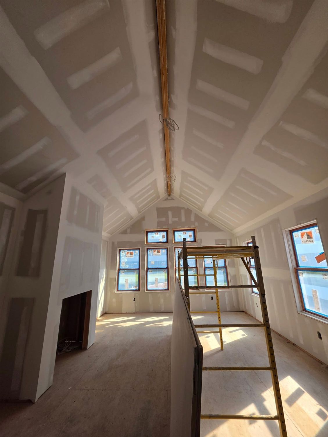 Interior of a house under construction. Drywall covers walls and angled ceiling. Scaffolding is set up near windows.
