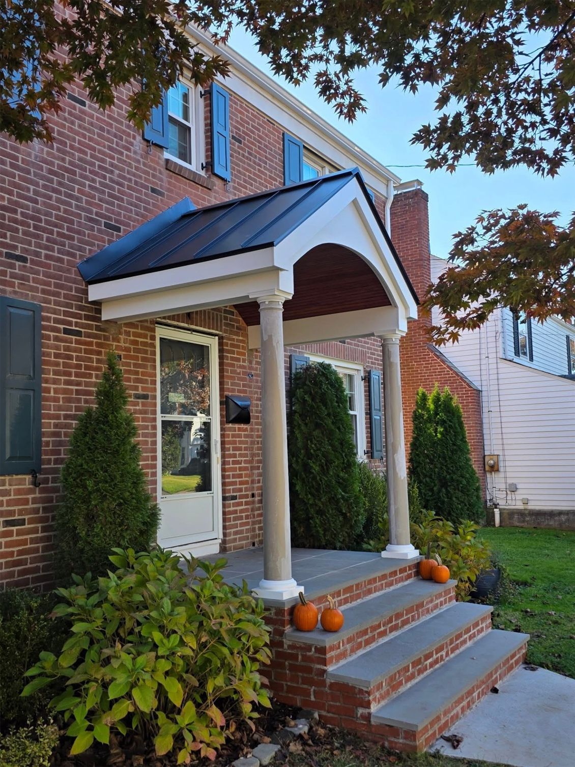 Brick house with a small front porch, gray steps, and pumpkins. Green shrubbery and blue shutters.